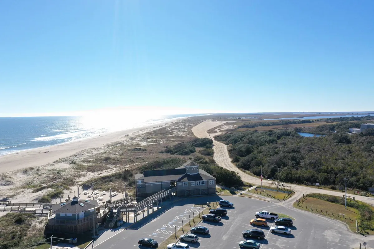 A wide exterior view of the beach with gentle waves, a visible boardwalk, parking lot, and lush coastal vegetation. The scene is active, with plenty of inviting space for sandcastle building, tide-pooling, and family beach fun.