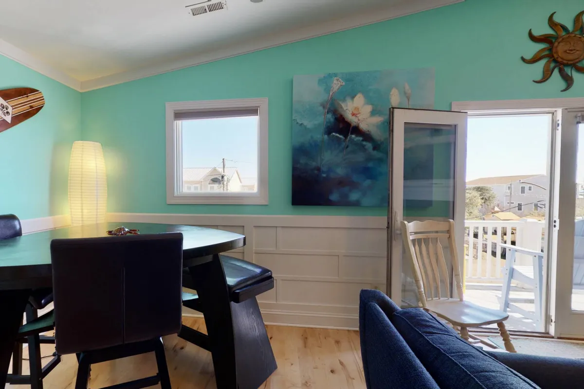 A cheerful dining area featuring a round table set for family meals, an abstract artwork on the wall, a large window letting in daylight, and a white rocking chair for relaxation. The setup feels intimate yet spacious, ideal for both memorable gatherings and quiet moments.