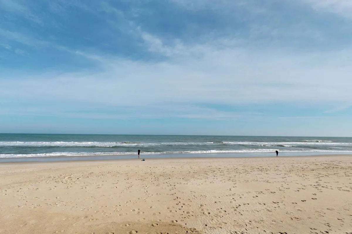 A serene exterior shot of a sandy beach at sunset, with gentle ocean waves and two figures walking along the shoreline. The tranquil setting captures the experience of evening strolls, beachcombing, and unwinding at the water's edge.