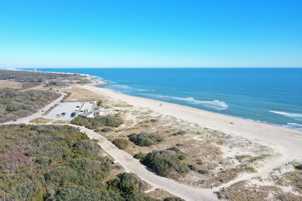 A bright, open exterior of the sandy beach and ocean framed by dunes and a nearby parking area. The setting suggests effortless transitions from car to sand, ideal for families with kids and lots of beach toys.