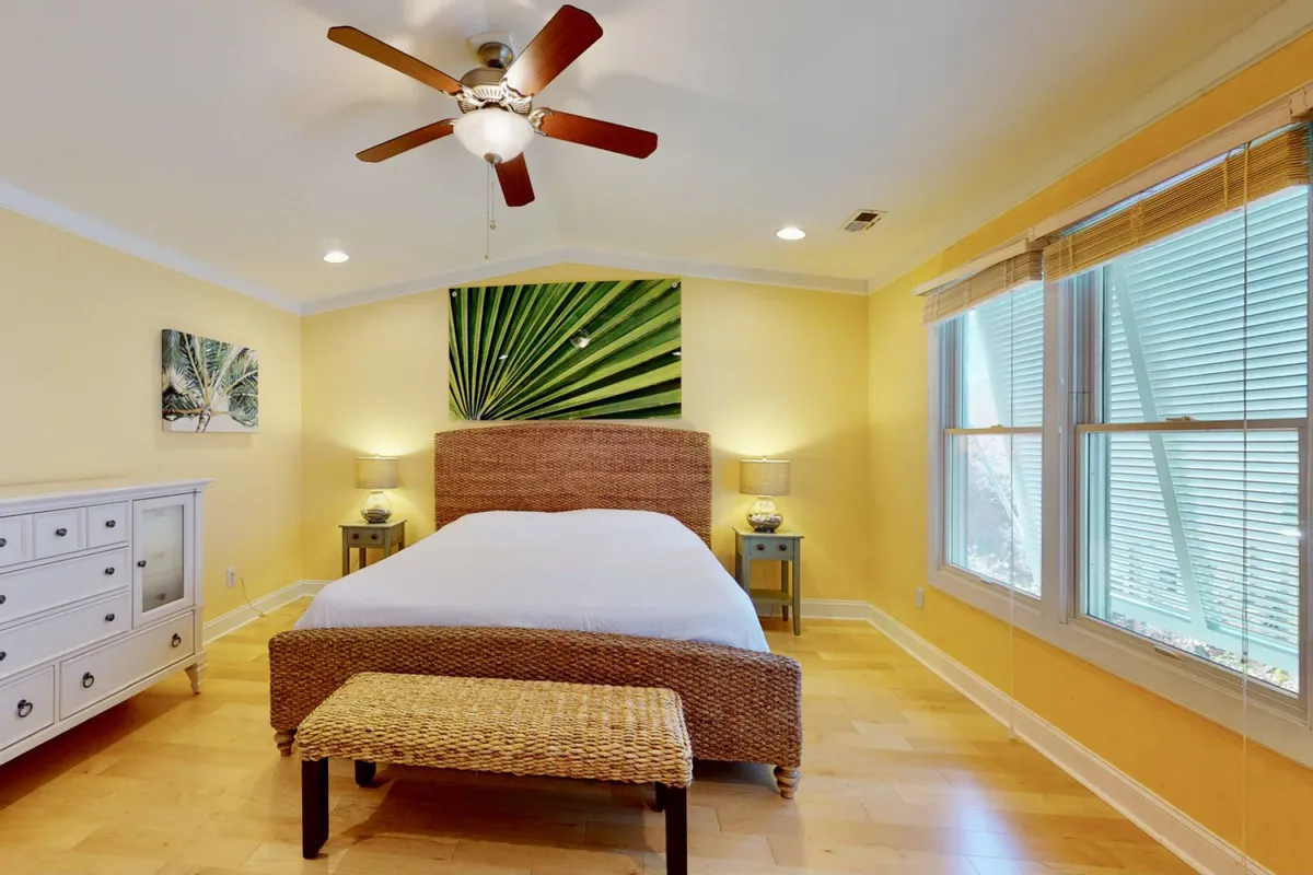 A serene bedroom with a woven bed frame, green leaf artwork above the bed, matching nightstands with warm lamps, and light-colored walls. The room exudes a tranquil, retreat-like atmosphere ideal for rest and relaxation.