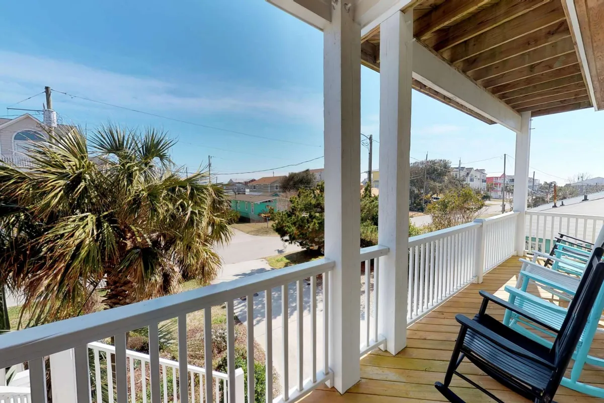 A covered balcony with classic rocking chairs overlooking palm trees and a quiet street, framed by a sturdy railing. The space is perfect for sipping morning coffee or enjoying the tranquil sounds of the ocean breeze, blurring the line between indoor comfort and outdoor beauty.