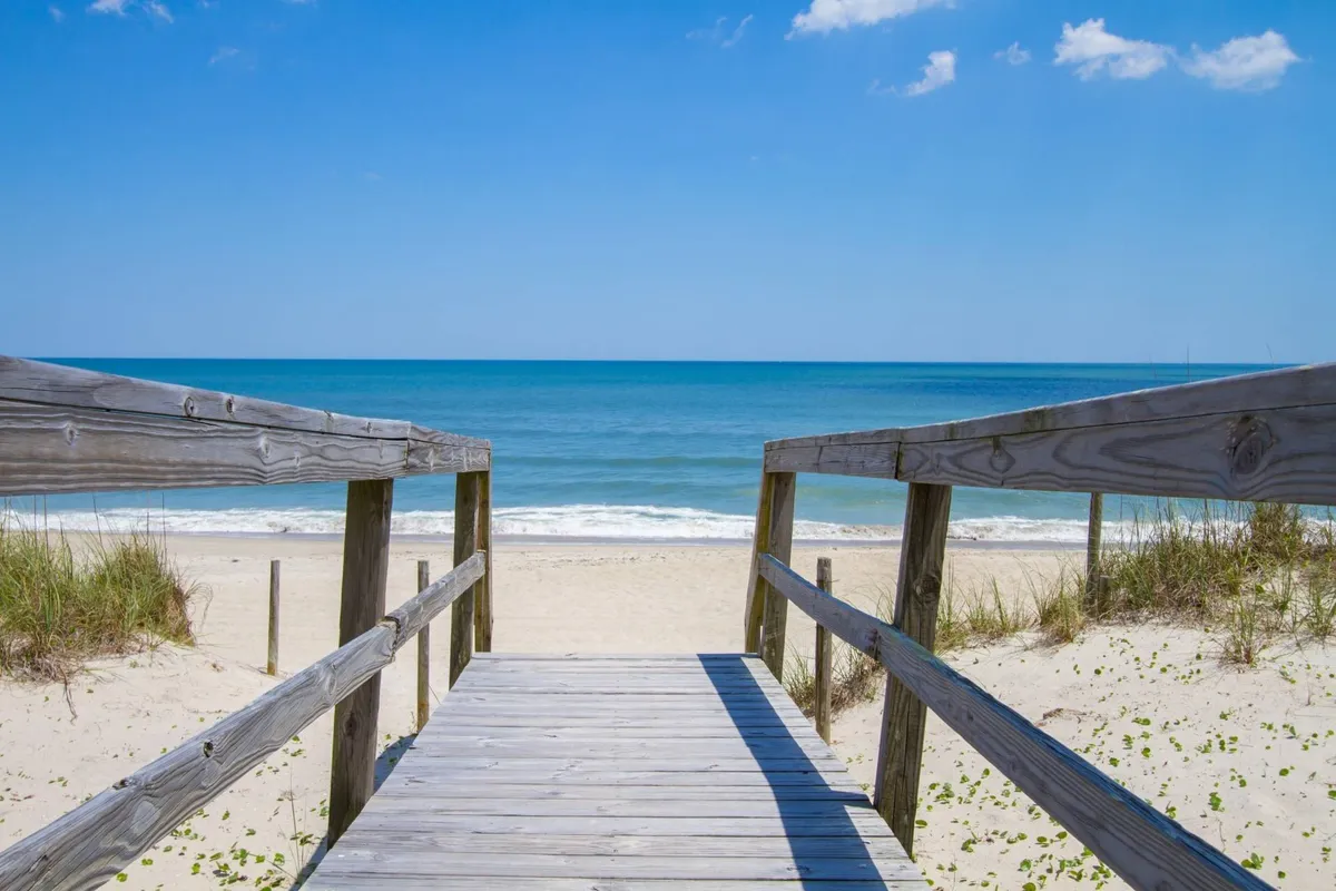 A wooden walkway leads over sand dunes toward the ocean, surrounded by dune grass and a sandy beach. The inviting path hints at easy access to the shore and plenty of room for walks, exploration, and outdoor adventures with pets.