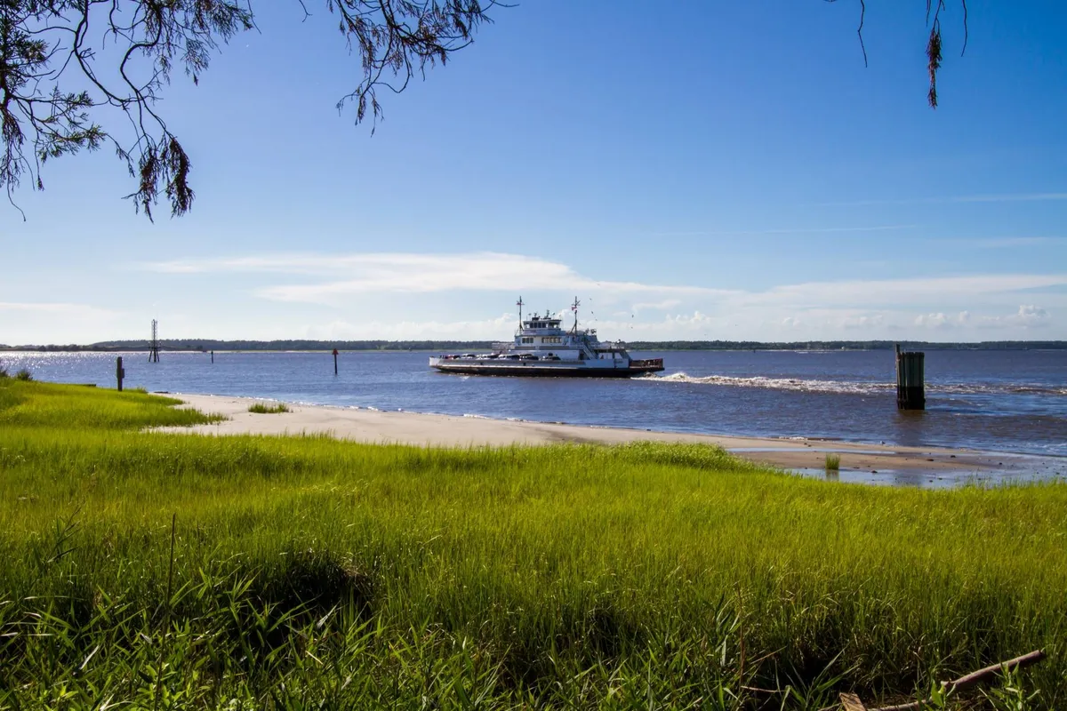 A scenic view of a ferry traveling across calm water, bordered by lush green grass under a bright blue sky. The landscape suggests peacefulness and the promise of exploration, evoking the experience of taking the Southport-Fort Fisher Ferry for a day of adventure.