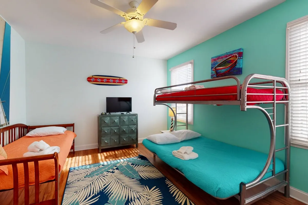 A playful and inviting bunk room with a bunk bed dressed in orange and turquoise bedding, vibrant wall art, a tropical rug, and a flat-screen TV. The space is designed for fun sleepovers and family comfort.