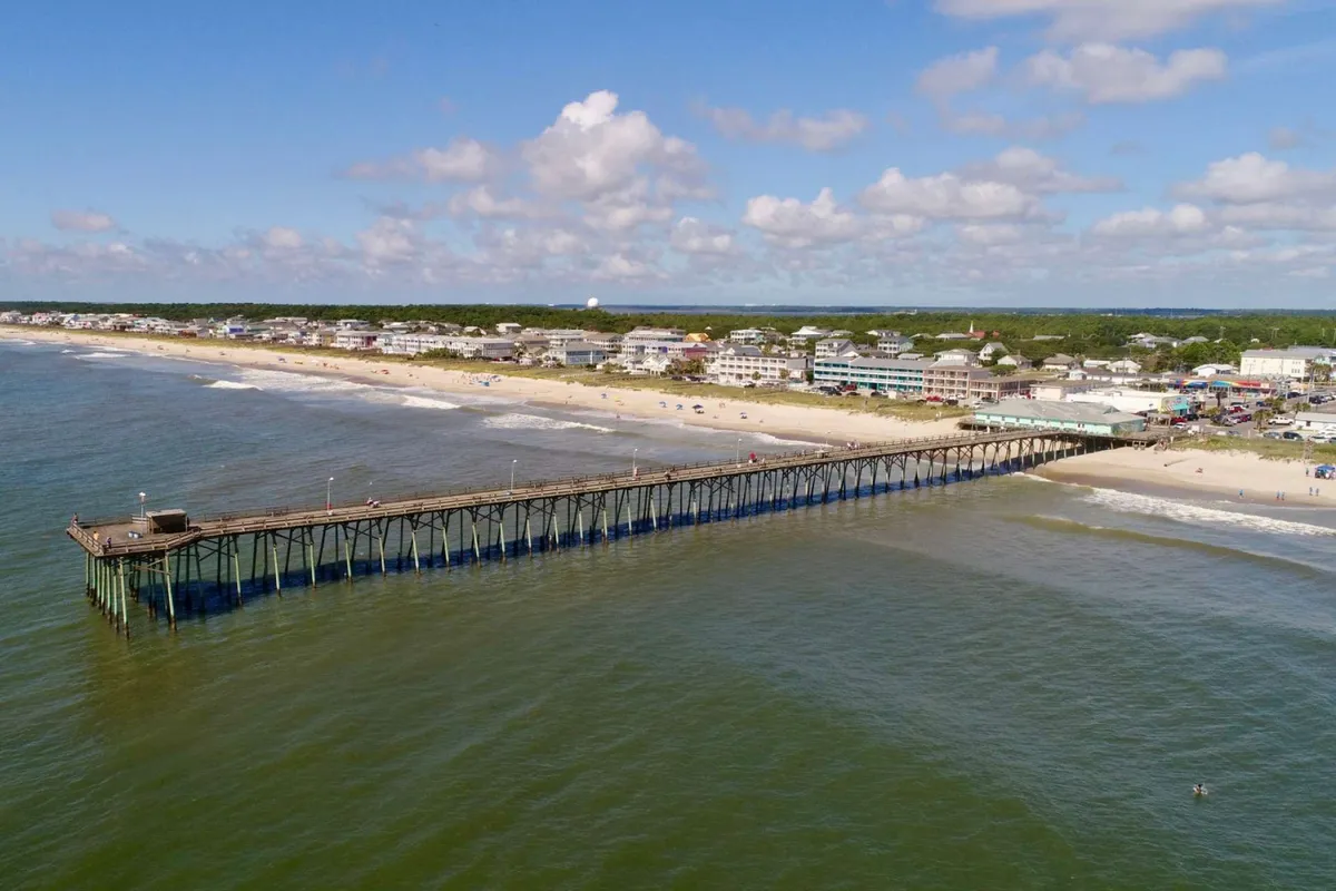 A scenic view featuring the Kure Beach Pier stretching into the ocean, with beachfront houses nestled alongside the sandy shoreline. People are visible fishing off the pier, sunbathing, and strolling along the water's edge. The sky is dotted with soft clouds, contributing to a relaxed, beach-day atmosphere.