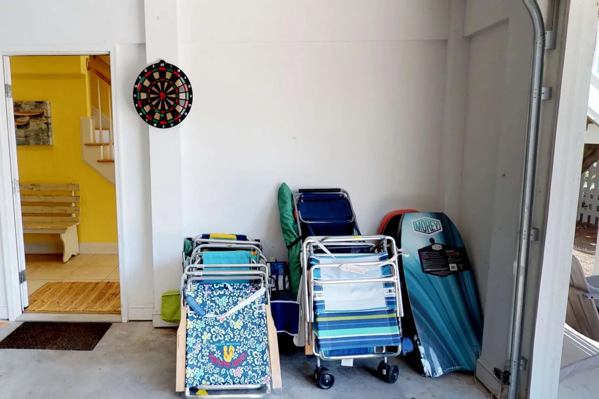 A playful indoor setup with a dartboard mounted on the wall, a stack of beach chairs ready for outings, and a surfboard adding a coastal flair. The area feels casual and multipurpose, hinting at beach adventures and laid-back indoor fun.