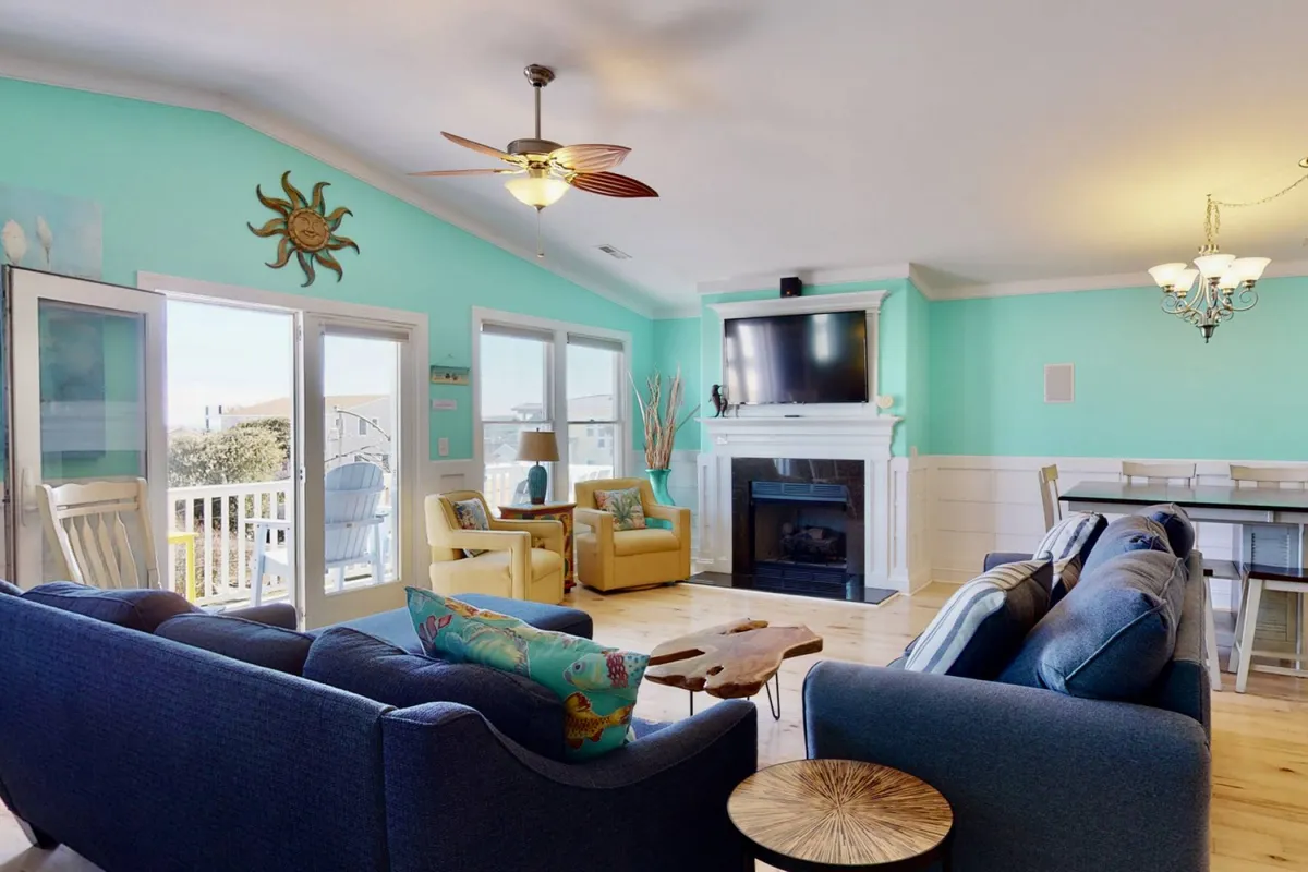 A spacious living room with bright turquoise walls, comfortable seating arranged for group gatherings, a fireplace for cozy evenings, large windows letting in natural light, and a dining area visible in the background. The space radiates warmth and togetherness, with ample room for relaxing, socializing, and enjoying games or movies.