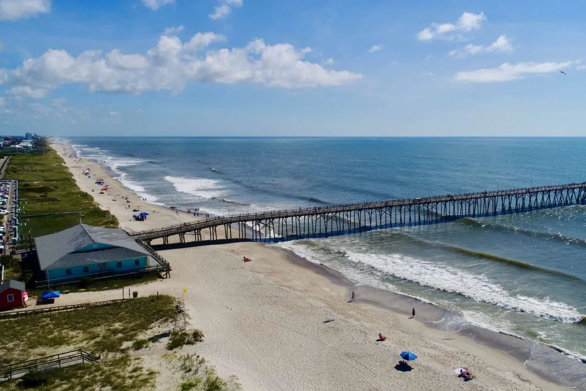 A serene view of a long pier stretching into the ocean, with families and children visible on the sand building sandcastles and enjoying the shoreline. Sunlight and clouds create a peaceful, playful mood.