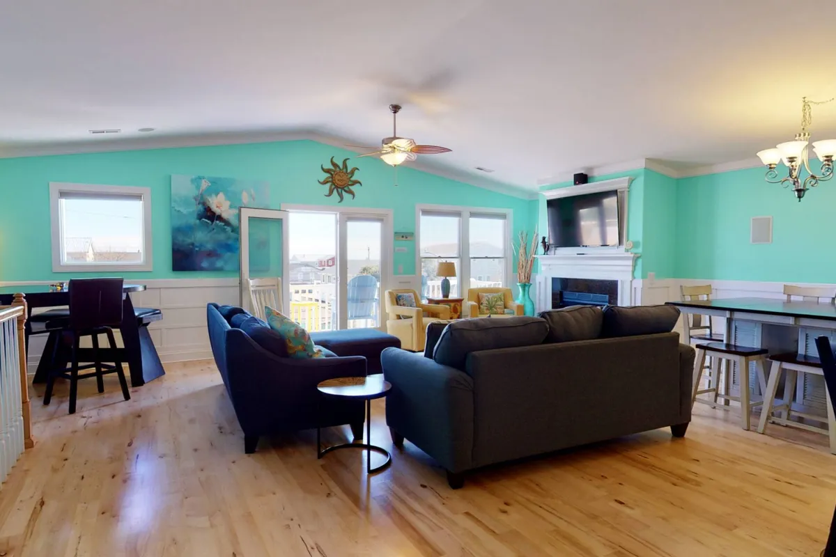 A bright, colorful living room with turquoise walls, wooden floors, and large windows, perfect for socializing or morning coffee. The layout supports gatherings, with decorative art and natural light creating a welcoming vibe for group activities or quiet retreats.