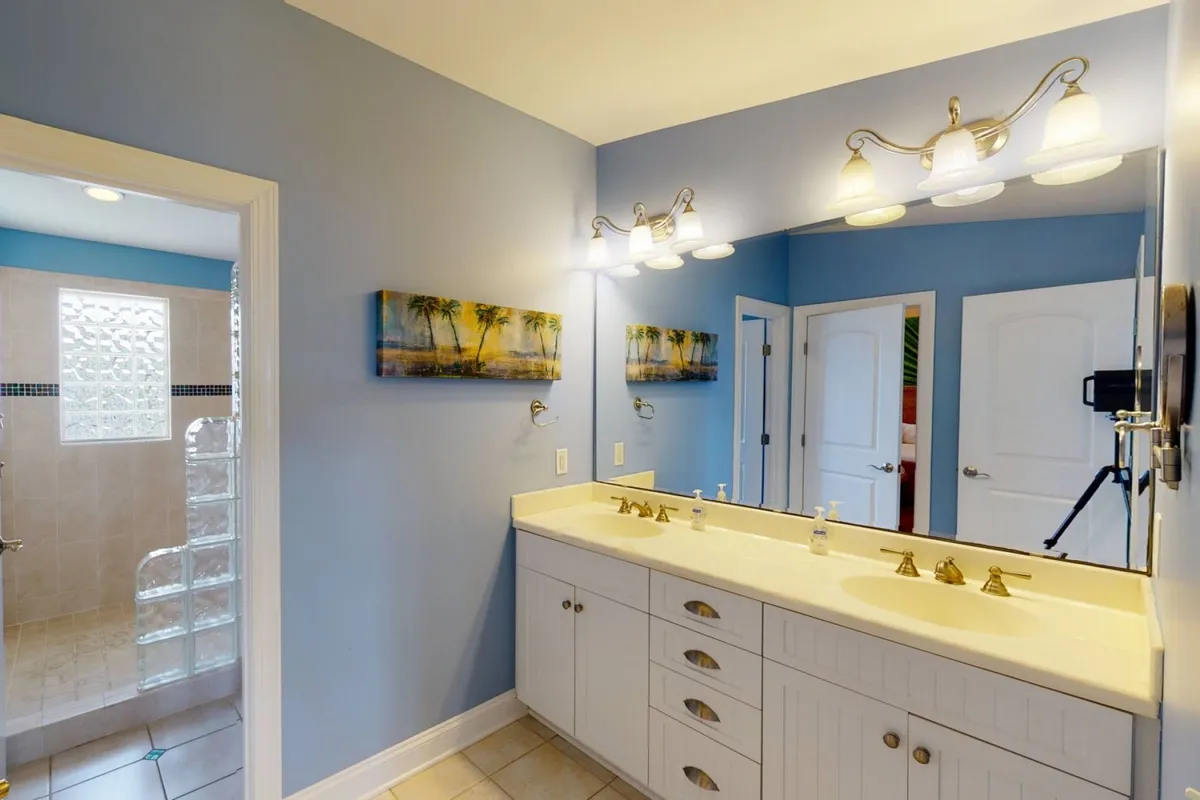A bright, serene bathroom featuring a dual sink vanity, framed artwork above, and a glass shower enclosure. The space is modern, clean, and ready for pampering routines.