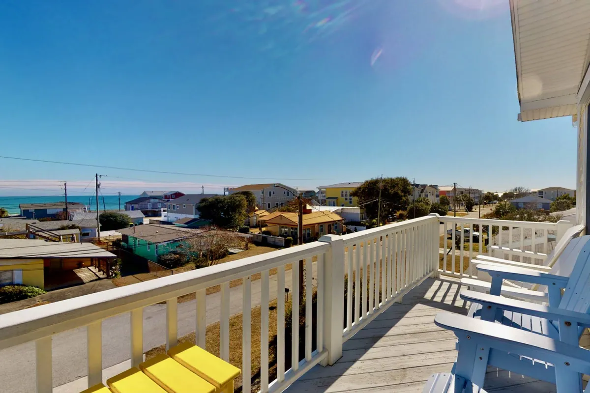 The exterior of a spacious townhome with a welcoming porch featuring bright yellow chairs overlooking a serene ocean view, neighboring beach houses, and a sun-drenched deck perfect for relaxing or socializing. The scene exudes a warm, inviting seaside atmosphere ideal for gatherings and unwinding just steps from the shore.