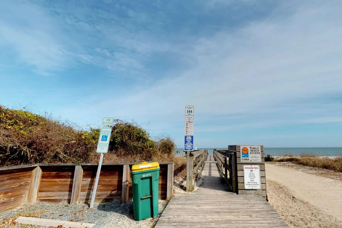 A wooden walkway leads toward public beach access, clearly marked by a beach access sign and flanked by greenery. The path is inviting and practical, illustrating the easy, direct route from the home to the sand—emphasizing the property’s proximity to the beach.